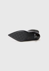 Black high-heeled shoe featuring a pointed toe, glossy upper, textured black sole with houndstooth pattern, and decorative accents.
