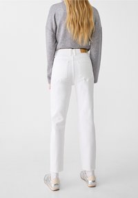 White high-waisted jeans with straight legs, featuring back pockets and a brown leather patch at the waistband, paired with a gray sweater.