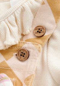 Tissu côtelé crème avec un col à volants, présentant un motif à carreaux jaunes et blancs et deux boutons décoratifs en bois.