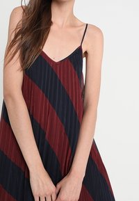 Ärmlös klänning i randig burgundy och marinblå. Tillverkad av lättviktsmaterial med en plisserad textur. Har tunna spagettiband och en avslappnad passform.