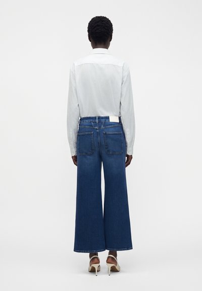 Personne aux cheveux courts et bouclés portant une chemise blanche à boutons, un jean bleu foncé à jambes larges et des talons blancs, debout de dos sur un fond uni.