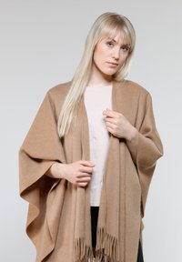 Beige gestrickter Poncho mit fransigen Kanten, weicher Textur, offenem Design und capeähnlichen Ärmeln, getragen über einem hellrosa Oberteil.