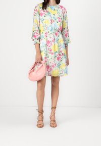 La robe fleurie présente un imprimé coloré de rose, jaune et vert sur un fond clair, avec des manches longues et une taille élastique. Associée à un sac à main rose et des sandales nude à lanières.