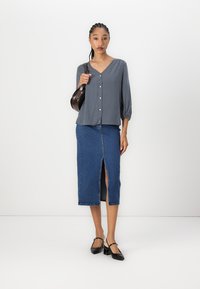 Marinebluse mit weißen Punkten, Knopfverschluss vorne; blaue Jeansrock mit vorderem Schlitz; schwarze Handtasche; schwarze Absatzschuhe.