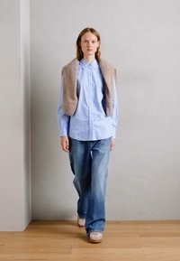 Chemise boutonnée bleu clair associée à un jean en denim délavé et ample. Un gilet texturé beige repose sur les épaules. Le mannequin marche sur un sol en bois.