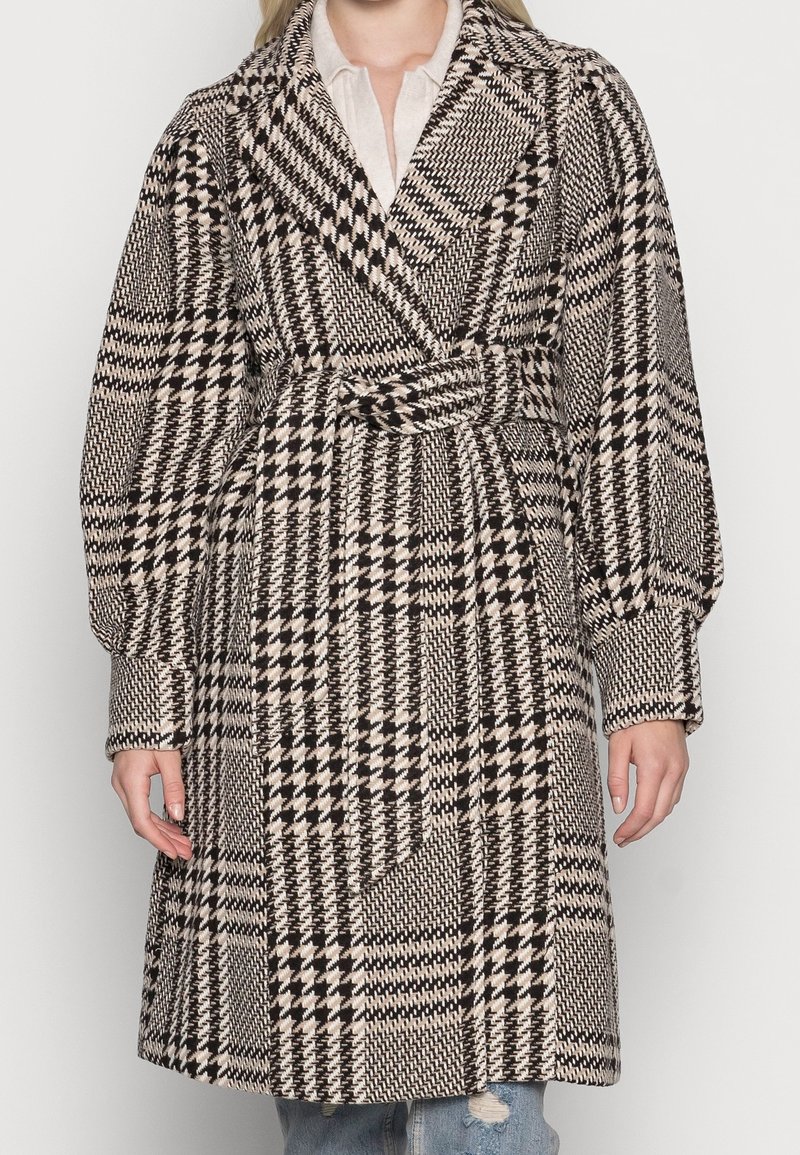 Femme portant un manteau ceinturé à motif pied-de-poule avec de larges revers et des manches bouffantes sur un jean déchiré.