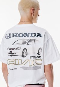 Witte oversized t-shirt met een grafisch ontwerp van een Honda Civic Type R, met grote zwarte en gouden tekst op de achterkant en een illustratie van de auto.