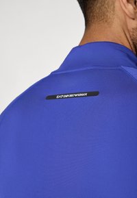 Plava sportska majica s glatkom teksturom, s crnom horizontalnom etiketom na stražnjem dijelu koja prikazuje "EA7 EMPORIO ARMANI" u bijelom tekstu.