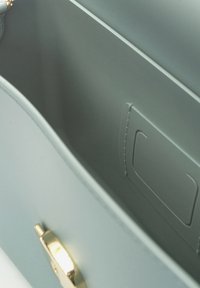 Interno della borsa di colore grigio chiaro con texture in pelle liscia, dotato di una chiusura in metallo dorato e una tasca rettangolare su un lato.