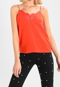 Top rojo sin mangas con encaje en el escote; tirantes finos y ajustables; combinado con jeans negros que tienen acentos de perlas blancas.