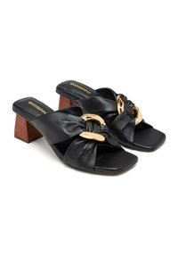 Des sandales en cuir noir à enfiler avec un bout carré, présentant un design noué et un accent en métal doré. Le talon est un bloc marron texturé.