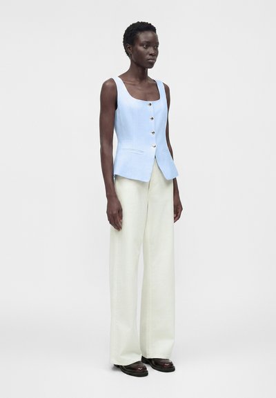 Γυναίκα που φοράει ανοιχτό γαλάζιο αμάνικο γιλέκο με κουμπιά, λευκό παντελόνι με φαρδιά μπατζάκια και σκούρα παπούτσια, στέκεται μπροστά σε απλό γκρι φόντο.