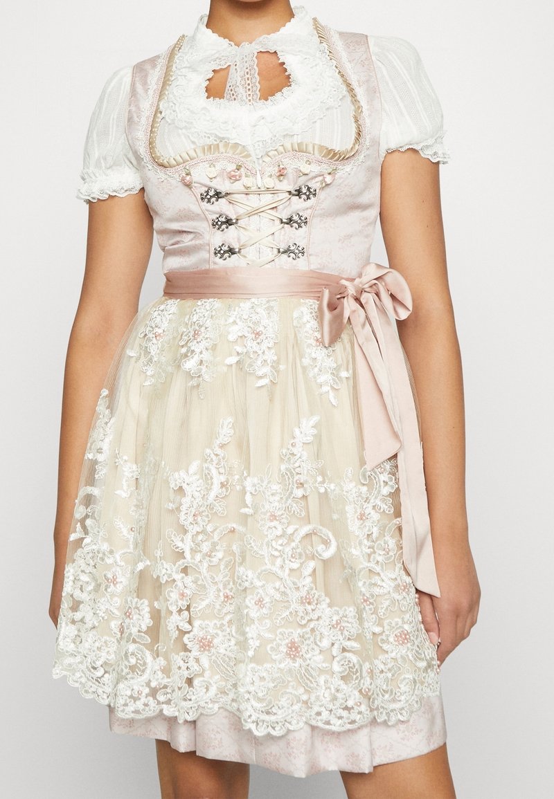 Femme portant une robe dirndl rose pâle avec un corsage à lacets, des manches ballons blanches et un tablier en dentelle crème noué avec un nœud en satin à la taille.