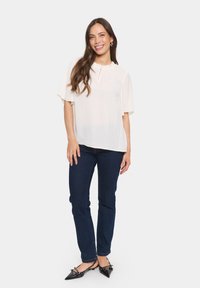 Ljus, krämfärgad blus med korta, svävande ärmar, hög halsringning och subtila veck. Matcher med mörkblå jeans och spetsiga svarta skor.