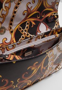 Borsa a tracolla nera e oro con motivo testurizzato e tasca interna con zip. Presenta un elegante design a catena su materiale liscio. L'etichetta reca la scritta "GUESS."