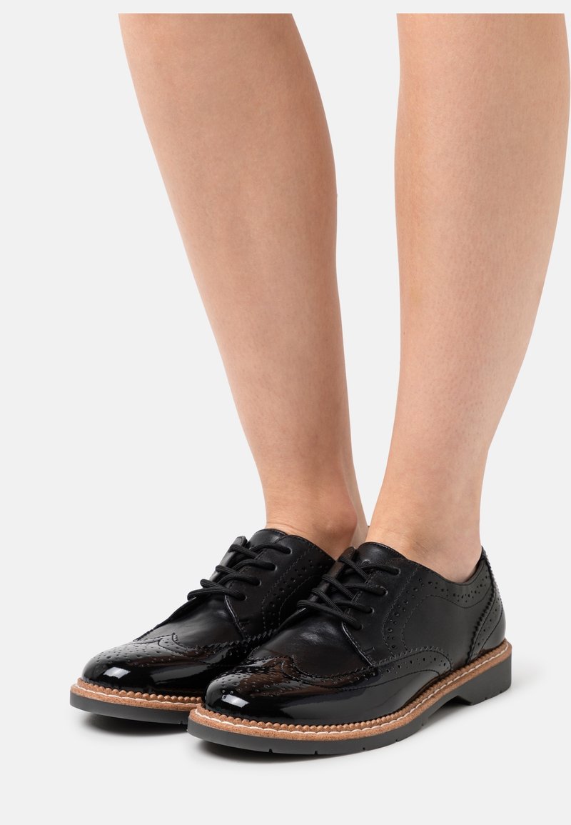 Zapatos brogue de cuero negro con acabado brillante, diseño de cordones, detalles perforados y suela de color marrón claro contrastante.