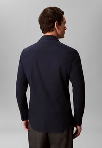 Mann, von hinten, trägt ein dunkelblaues, langärmeliges Hemd mit Knöpfen und dunkle Hosen vor einem schlichten grauen Hintergrund.