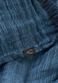 Écharpe bleue tissée avec un motif texturé, ornée de bords frangés et d'une petite étiquette noire avec un logo. Matériau doux et design de poids moyen.