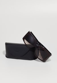 Gafas de sol de carey marrón con lentes oscuros, reposando sobre un estuche de cuero negro grabado con "GUESS". El estuche tiene una tapa redondeada.