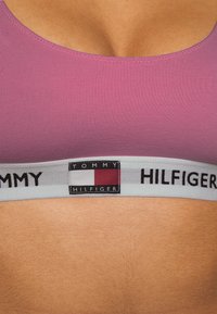 Bralette púrpura sin costuras con una banda elástica gris contrastante que presenta el logo "TOMMY HILFIGER". Textura suave, escote redondo y ajuste ceñido.