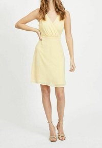 Vestido amarillo sin mangas con un diseño envolvente, de longitud hasta la rodilla y textura fluida. Combinado con sandalias ligeras de tiras.