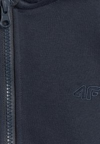 Gros plan d'un tissu bleu foncé texturé avec une fermeture éclair partiellement visible à gauche et un logo brodé "4F" à droite.
