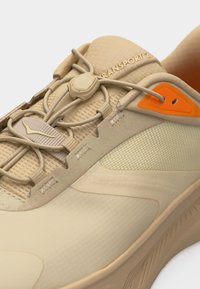 Μπεζ αθλητικό παπούτσι με ελαστικούς κορδόνια με ρυθμιστικό, επάνω μέρος από διχτυωτό ύφασμα, πορτοκαλί λεπτομέρεια γύρω από τις θηλιές και το κείμενο "TRANSPORT 2" στη μαλακή γλώσσα.