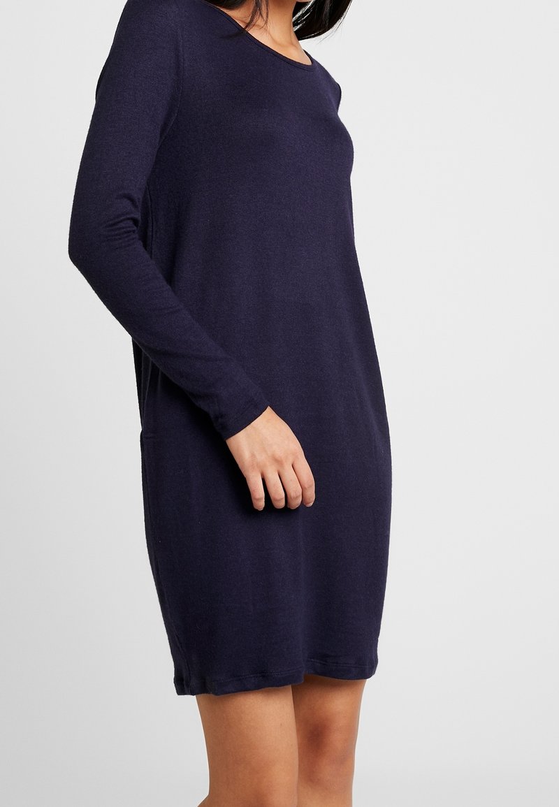 Femme portant une robe bleu marine à manches longues, longueur genoux, debout devant un fond uni gris clair.