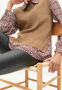 Braunes gestricktes ärmelloses Oberteil über einer gemusterten (kastanienbraunen und weißen) Bluse, kombiniert mit schwarzen, eng anliegenden Hosen. Modell sitzt auf einem Holzstuhl.