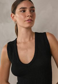 Svart ärmlös topp med djup V-ringning, texturerad tyg och ribbad nederkant. Modellen har minimal sminkning och små hoops. 