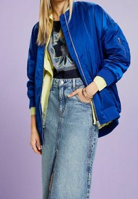 Personne portant une veste bomber bleue par-dessus une chemise jaune et un tee-shirt graphique noir, assortie d'une jupe en denim taille haute avec une fente sur le devant.