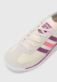Baskets de sport beiges avec des accents violets et roses, dotées d'une languette texturée, de lacets ronds et d'une semelle en caoutchouc blanc.
