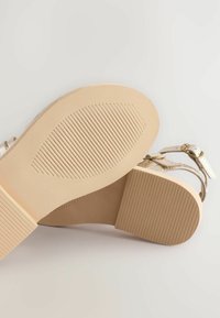 Beżowe sandały z gładką, teksturowaną podeszwą. Oferują regulowane paski wokół kostki i płaską podstawę dla stabilności. Prostota designu bez ozdobników.