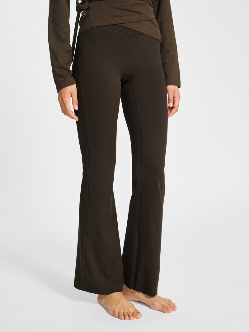 Person, die dunkelbraune ausgestellte Hose und ein passendes Oberteil trägt, steht barfuß auf hellem Hintergrund.