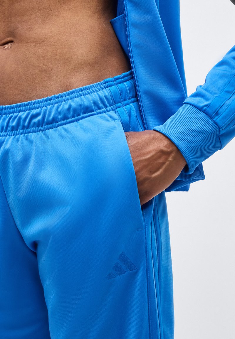 Primo piano di una persona che indossa pantaloni e giacca sportivi blu, con la mano in tasca e l'addome tonico visibile.