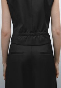 Vista posteriore di una persona che indossa un top nero aderente senza maniche con dettagli elasticizzati e pantaloni neri abbinati con tasche posteriori orizzontali.