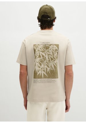 Mężczyzna noszący beżowy t-shirt z zielonym nadrukiem bambusa i napisem na plecach, w połączeniu z zieloną czapką i kremowymi spodniami, zwrócony tyłem.