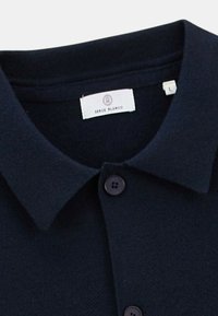 Polo de couleur bleu marine en tissu texturé avec un col classique, deux boutons noirs et une étiquette indiquant "Serge Blanco" et la taille "L".