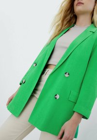 Femme portant un blazer croisé vert vif sur un crop top côtelé beige et un pantalon beige clair, debout devant un fond clair uni.