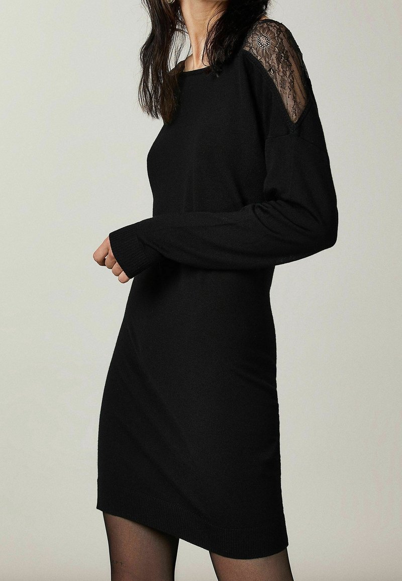 Femme portant une robe en maille noire à manches longues avec des détails en dentelle transparente aux épaules, debout devant un fond clair uni.