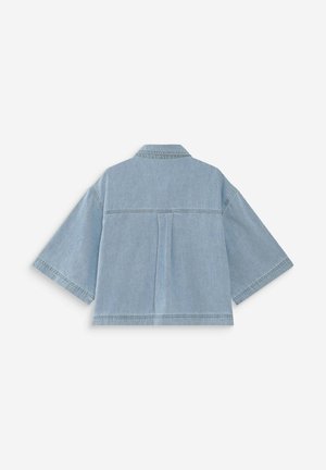 Camisa corta de mezclilla azul claro con mangas anchas de medio largo y un pliegue central en la espalda sobre un fondo blanco liso.