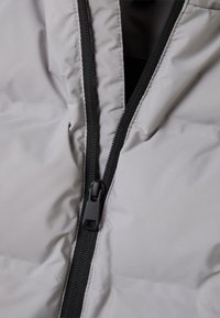 Chaqueta aislante de color gris claro con cremallera frontal, que presenta una pista de cremallera negra y un tirador. Textura suave, material ligero.