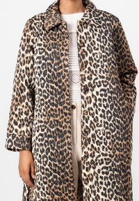 Femme portant un manteau matelassé à imprimé léopard boutonné jusqu'aux genoux, sur un haut blanc en dentelle et plissé, associé à un pantalon beige taille haute.