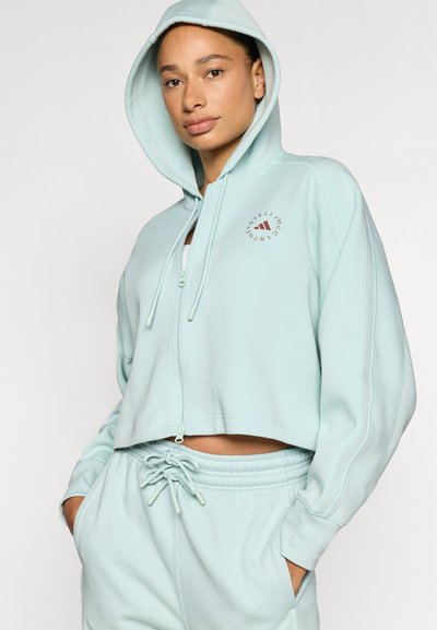 Κοντή φούτερ με κουκούλα σε μέντα πράσινο χρώμα, με φερμουάρ μπροστά, κορδόνι στην κουκούλα και λογότυπο στην αριστερή πλευρά. Μαλακό, λείο ύφασμα με ριμπ μανσέτες.