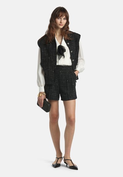 Femme portant un gilet en tweed noir avec un short assorti, une blouse blanche avec un nœud noir, une petite pochette noire, un bracelet en or et des escarpins noirs à petits talons.