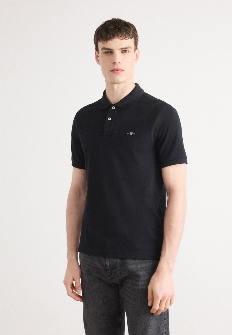 Black polo shirt with a classic collar, two buttons, and an embroidered logo on the left chest. Cotton material; short sleeves, minimal design.
