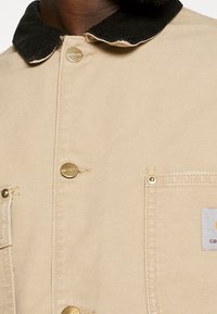 Beige lærredjakke med en sort kordfløjlskrave, guldfarvede knapper og en brystlomme med et logo-patch. Glat tekstur med en struktureret design.