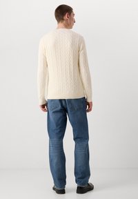 Maglione crema in maglia con motivo a trecce, polsini e orlo a coste, abbinato a jeans blu a gamba dritta e scarpe nere.