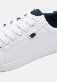 Zapatillas de lona en blanco con superficie texturizada, que presentan detalles en azul marino, cordones redondos y una pequeña etiqueta roja y azul en el lateral.