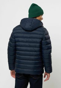 Marine blauwe pufferjack met horizontale quiltpatronen, een capuchon en elastische manchetten, gecombineerd met een groene gebreide beanie.